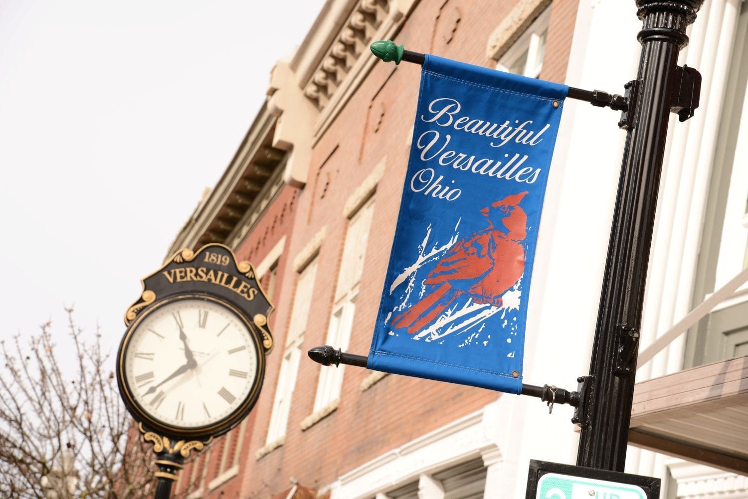 A blue downtown banner reading “Beautiful Versailles Ohio” with a red cardinal illustration hangs from a black lamppost in front of historic brick storefronts. An ornamental street clock marked “1819 Versailles” stands nearby, highlighting the community’s small-town streetscape.