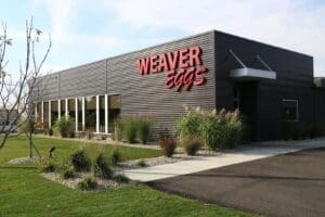 Exterior view of the Weaver Eggs headquarters, a modern dark-gray metal building with large red “WEAVER Eggs” signage mounted on the front. Landscaping with green lawns, ornamental grasses, shrubs, and a concrete walkway surrounds the facility under a bright, partly cloudy sky.